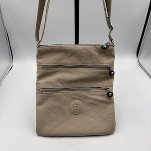 May include: A tan crossbody bag with three zippered compartments and a long adjustable strap. The bag features a circular logo patch on the front and silver-toned hardware. The bag is made of a lightweight, durable material.