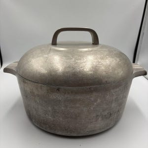 May include: A round, brushed aluminum Dutch oven with a matching lid. The pot features two integrated side handles and a central arched handle on the lid. The metal surface shows a matte, weathered texture with visible signs of use and wear.