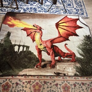 May include: A rectangular tapestry featuring a red dragon exhaling fire. The dragon has large wings and is positioned on a stone surface. The background depicts a castle and trees. The tapestry has a brown border.