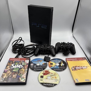 May include: A black PlayStation 2 console standing vertically with two black wired controllers, two power and AV cables, and five game discs. The collection includes The Sims 2 and Crazy Taxi game cases, plus three loose PlayStation 2 game discs.