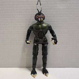 May include: A black and green action figure of an insectoid alien with a segmented body, insect-like head, and clawed feet. The figure has a metallic sheen on its chest and head, with orange and black details. It stands upright with arms at its sides.