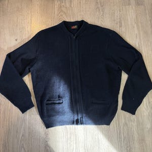 May include: Navy blue ribbed knit zip-up cardigan sweater with two front pockets. The sweater has a classic design with a ribbed texture and a full-length zipper. The collar is a crew neck.