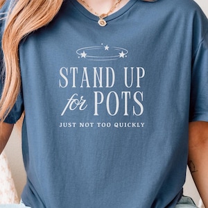 Puede incluir: Una camiseta azul con el texto blanco "STAND UP for POTS", con la frase "JUST NOT TOO QUICKLY" debajo. La camiseta tiene cuello redondo y mangas cortas.