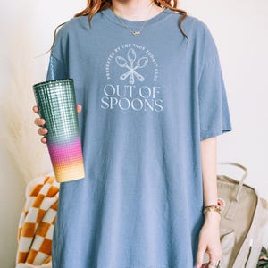 Pode incluir: Uma camiseta azul com o texto "OUT OF SPOONS" e um desenho de colher. A camisa &eacute; combinada com um copo alto e iridescente com canudo. O copo tem um gradiente de cores, incluindo rosa, laranja e amarelo.