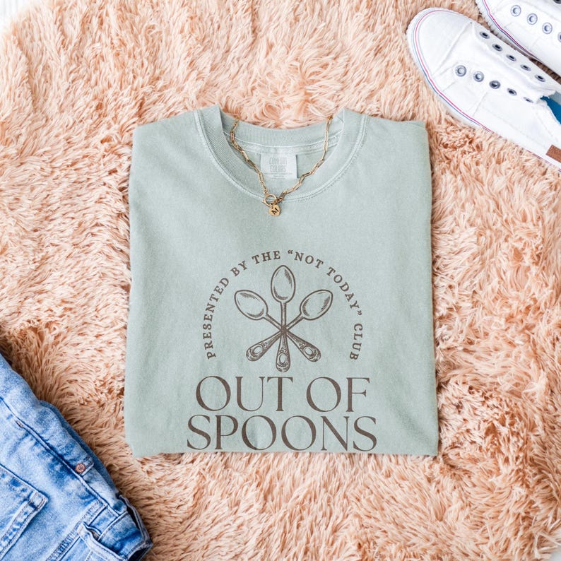 Pode incluir: Camisola verde claro com o texto "OUT OF SPOONS" e "PRESENTED BY THE 'NOT TODAY' CLUB" em castanho. O design inclui colheres cruzadas. Um colar de ouro e t&ecirc;nis brancos tamb&eacute;m s&atilde;o vis&iacute;veis.