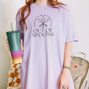 Pode incluir: Uma camiseta lil&aacute;s clara com o texto "OUT OF SPOONS" e um gr&aacute;fico de colher. A camiseta &eacute; apresentada por "THE NOT TODAY CLUB". Um copo alto e colorido com canudo &eacute; segurado na m&atilde;o esquerda.