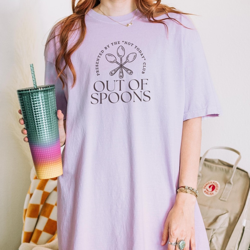 Pode incluir: Uma camiseta lil&aacute;s clara com o texto "OUT OF SPOONS" e um gr&aacute;fico de colher. A camiseta &eacute; apresentada por "THE NOT TODAY CLUB". Um copo alto e colorido com canudo &eacute; segurado na m&atilde;o esquerda.
