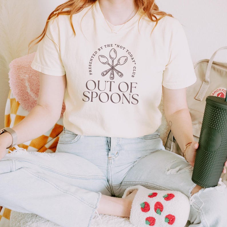 Pode incluir: Camiseta creme com o texto "OUT OF SPOONS" e a frase "PRESENTED BY THE 'NOT TODAY' CLUB" em um design circular. A camiseta &eacute; combinada com jeans claros e chinelos de morango. Um copo verde &eacute; segurado.