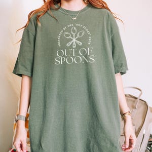 Pode incluir: Camiseta verde-oliva com o texto branco "OUT OF SPOONS" e um gr&aacute;fico de colher. A camisa tamb&eacute;m apresenta o texto "PRESENTED BY THE 'NOT TODAY' CLUB" em um design circular ao redor do gr&aacute;fico da colher.