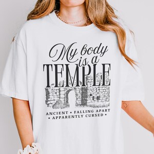 May include: White t-shirt with the text "My body is a TEMPLE" in a black, elegant font. Below the text is a graphic of a stone structure with an arched doorway. The phrases "ANCIENT", "FALLING APART", and "APPARENTLY CURSED" are also printed.