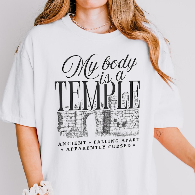 May include: White t-shirt with the text "My body is a TEMPLE" in a black, elegant font. Below the text is a graphic of a stone structure with an arched doorway. The phrases "ANCIENT", "FALLING APART", and "APPARENTLY CURSED" are also printed.
