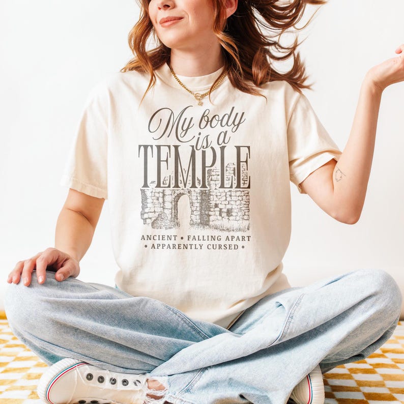 May include: Cream-colored t-shirt with the text "My body is a TEMPLE" in a stylized font, with an illustration of an ancient structure. Below the text are the words "ANCIENT &bull; FALLING APART &bull; APPARENTLY CURSED."