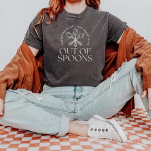 Pode incluir: T-shirt cinza escuro com o texto "OUT OF SPOONS" e a frase "PRESENTED BY THE 'NOT TODAY' CLUB" em um design circular. A camisa &eacute; usada com jeans azul claro e t&ecirc;nis brancos.