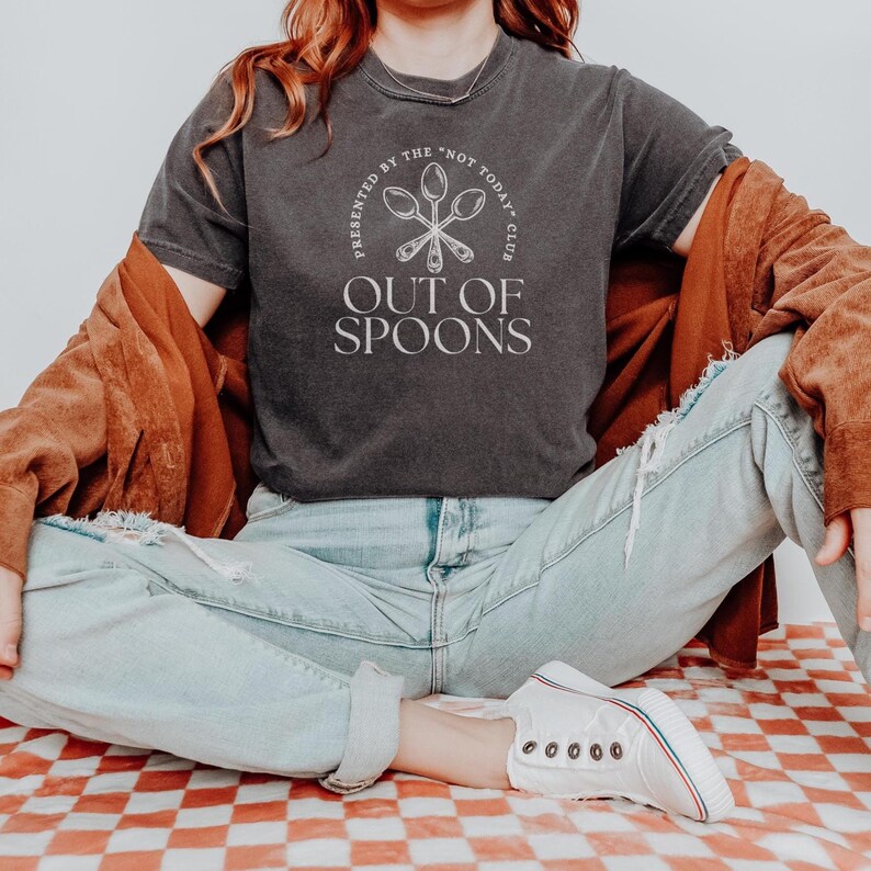 Pode incluir: T-shirt cinza escuro com o texto "OUT OF SPOONS" e a frase "PRESENTED BY THE 'NOT TODAY' CLUB" em um design circular. A camisa &eacute; usada com jeans azul claro e t&ecirc;nis brancos.