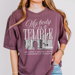 May include: A mauve-colored t-shirt with the text "My body is a TEMPLE" in white script and block letters. Below the text is a graphic of an ancient stone structure. The words "ANCIENT - FALLING APART - APPARENTLY CURSED" are below the graphic.