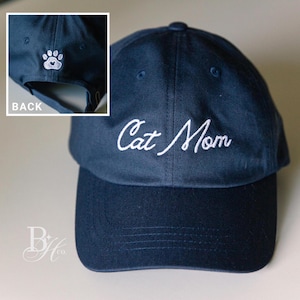 May include: Navy blue baseball cap with the embroidered text "Cat Mom" in white script. The back of the cap features a white paw print design. A stylish accessory for cat lovers.