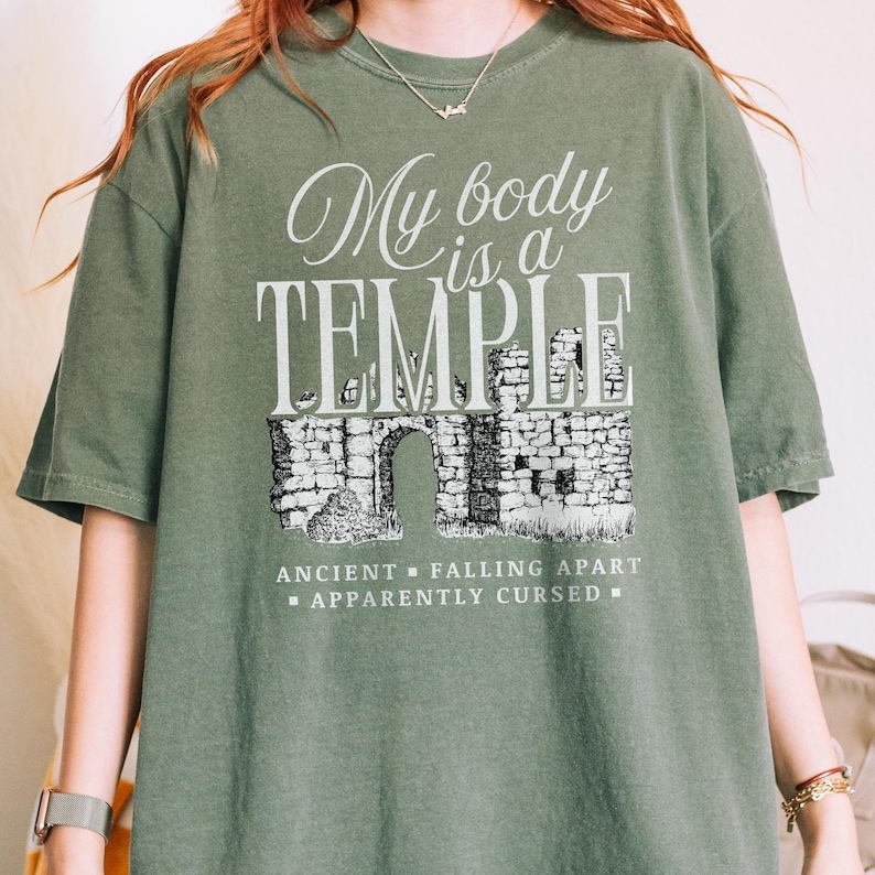 May include: An olive green t-shirt with the text "My body is a TEMPLE" in a decorative font. Below the text is a black and white illustration of an ancient stone structure with an arched doorway. The shirt also has the text "ANCIENT - FALLING APART - APPARENTLY CURSED".