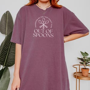 Pode incluir: Camiseta de cor malva com a frase branca &laquo; OUT OF SPOONS &raquo; sob um desenho circular com colheres e as palavras &laquo; PRESENTED BY THE 'NOT TODAY' CLUB &raquo;. A camiseta tem mangas curtas e um corte folgado.