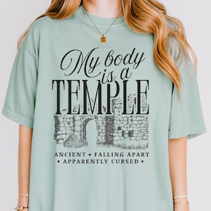 May include: A light green t-shirt with the text "My body is a TEMPLE" in black script and block letters. Below the text is a black and white illustration of an ancient stone structure with an arched doorway. The shirt also has the phrases "ANCIENT &bull; FALLING APART &bull; APPARENTLY CURSED."