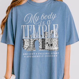 May include: A light blue t-shirt with the text "My body is a TEMPLE" in white script and block letters. Below the text is a black and white illustration of an ancient stone structure with an arched doorway. The text "ANCIENT - FALLING APART - APPARENTLY CURSED" is below the illustration.