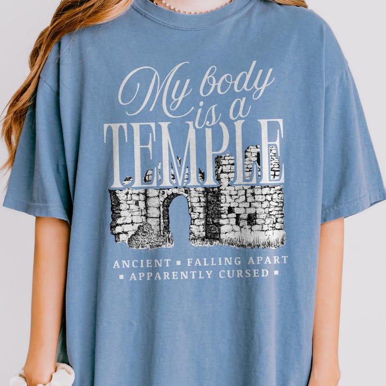 May include: A light blue t-shirt with the text "My body is a TEMPLE" in white script and block letters. Below the text is a black and white illustration of an ancient stone structure with an arched doorway. The text "ANCIENT - FALLING APART - APPARENTLY CURSED" is below the illustration.