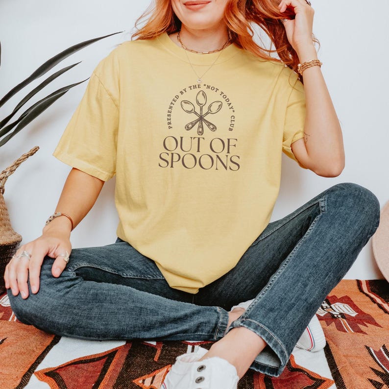 Pode incluir: Uma camiseta amarela com o texto "OUT OF SPOONS" em marrom, com um gr&aacute;fico de colher. A camisa tamb&eacute;m tem o texto "PRESENTED BY THE 'NOT TODAY' CLUB" em um design circular. A pessoa est&aacute; usando jeans azul.