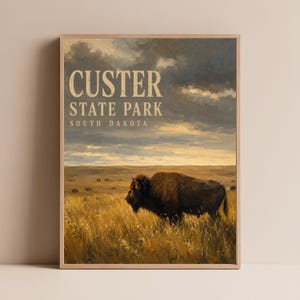 May include: A framed wall art print featuring an oil painting of a bison standing in a golden prairie field under a dramatic, cloudy sky. The text CUSTER STATE PARK SOUTH DAKOTA is printed in a light beige serif font in the upper portion of the image.