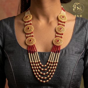 May include: A multi-strand necklace featuring gold-toned circular pendants with red accents and intricate detailing. The necklace incorporates red and cream-colored beads, creating a layered effect. The jewelry is displayed on a person wearing a dark gray top.