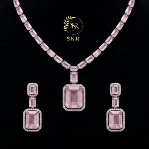 May include: A pink gemstone jewelry set, including a necklace and earrings, against a black background. The necklace and earrings feature rectangular pink stones with a white border. The necklace has a large pendant. The earrings are dangling.