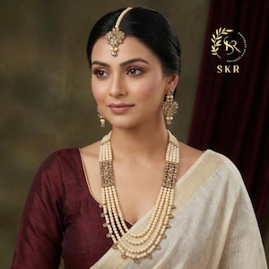 May include: A woman wearing a pearl necklace, earrings, and a headpiece. The necklace has multiple strands of pearls with ornate gold-colored rectangular clasps. The earrings and headpiece match the necklace design. The image includes the text "SKR".