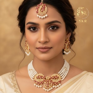 Może przedstawiać: Kobieta nosi złoty zestaw biżuterii składający się z naszyjnika, kolczyków i nakrycia głowy. Naszyjnik ma wiele sznurów białych pereł z centralną złotą zawieszką ozdobioną czerwonymi kamieniami. Kolczyki i nakrycie głowy odzwierciedlają wzór zawieszki.