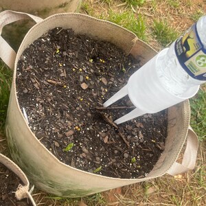 Pode incluir: Uma foto em close-up de um saco de cultivo de tecido bege cheio de terra escura. Um dispositivo de irriga&ccedil;&atilde;o de pl&aacute;stico transparente &eacute; inserido no solo, com uma garrafa de &aacute;gua presa. O saco tem duas al&ccedil;as e est&aacute; na grama.