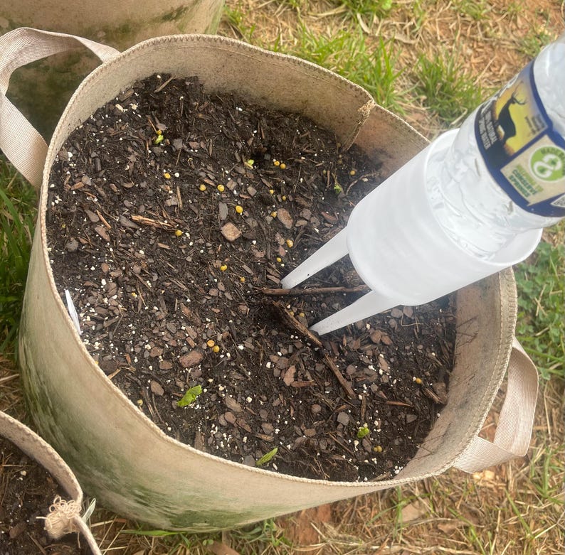 Pode incluir: Uma foto em close-up de um saco de cultivo de tecido bege cheio de terra escura. Um dispositivo de irriga&ccedil;&atilde;o de pl&aacute;stico transparente &eacute; inserido no solo, com uma garrafa de &aacute;gua presa. O saco tem duas al&ccedil;as e est&aacute; na grama.