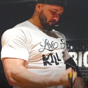 Könnte beinhalten: Ein weißes T-Shirt mit dem Text "Love Strong Kill It" in schwarzer Schrift und Blockbuchstaben. Die Person auf dem Bild trägt eine schwarze Kappe und einen schwarzen Armärmel. Der Hintergrund ist dunkel.