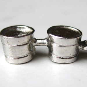 May include: A silver-toned charm featuring two small, connected buckets. Each bucket has a rounded top and a horizontal band around the middle. A small loop is attached to one side, suggesting it could be used as a pendant or charm.