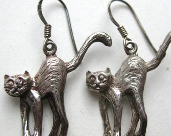 May include: A pair of silver-toned drop earrings featuring a cat design with an arched back and a raised tail. Each earring is suspended from a curved wire hook, showcasing a textured metallic finish that highlights the feline form and facial features.
