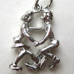 May include: A polished silver-toned metal charm featuring two figures facing each other while dancing. The figures are joined at the hands, creating a heart-shaped negative space between them. A small circular jump ring is attached to the top of the charm.