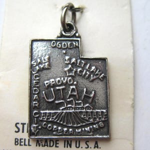 May include: A silver-coloured Utah state charm pendant. The charm is shaped like the state of Utah and features the names of cities like Ogden, Salt Lake City, and Provo. The text "BELL MADE IN U.S.A." is visible.