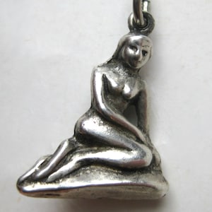 May include: A silver-toned metal charm depicting a figure in a seated, reclining pose. The figure has long hair and is positioned on a textured, rock-like base. A small circular jump ring is attached to the top of the figure's head for hanging.