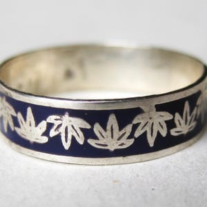 May include: A silver band ring featuring a continuous inlay of deep blue enamel. The surface of the ring is decorated with a repeating pattern of silver leaf motifs that are set against the dark blue background, creating a distinct contrast.