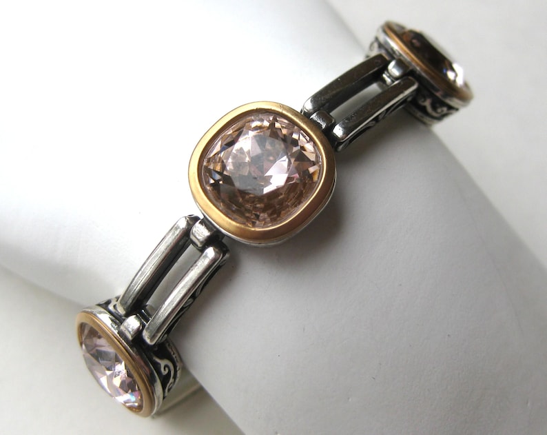 May include: A bracelet featuring three square-shaped, light champagne-colored faceted crystals set in gold-toned bezels. The links connecting the crystals are made of oxidized silver-toned metal with intricate scrollwork patterns on the sides.