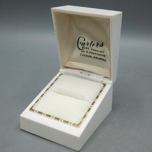 Op de afbeelding: Open witte sieradendoos met een crèmekleurig interieur en een goudkleurige rand. De binnenkant van het deksel heeft de tekst "Carters Fine Jewelry, 66 E. Pennington, Tucson, Arizona". De doos is ontworpen om een ring of andere kleine sieraden in te bewaren.