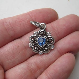 May include: A silver-toned metal pendant featuring a central heart-shaped blue gemstone surrounded by swirling filigree patterns and small clear stones. The pendant includes a twisted metal jump ring at the top for attachment to a chain or cord.
