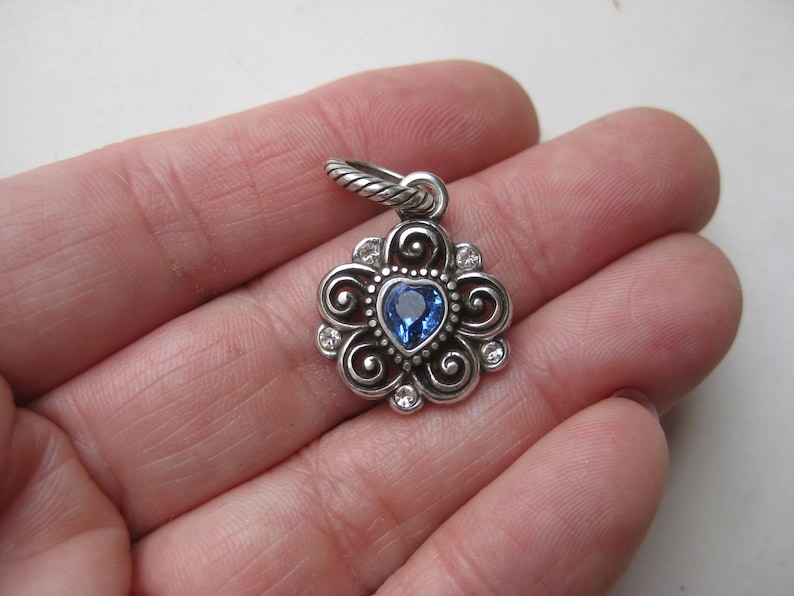 May include: A silver-toned metal pendant featuring a central heart-shaped blue gemstone surrounded by swirling filigree patterns and small clear stones. The pendant includes a twisted metal jump ring at the top for attachment to a chain or cord.
