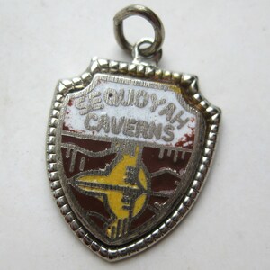 May include: A silver-toned shield-shaped charm featuring the text SEQUOYAH CAVERNS in raised silver letters against a white background. The lower portion displays a brown and yellow graphic design with a central yellow shape and a small jump ring.