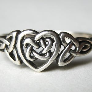 May include: A silver ring featuring a Celtic knot heart design. The ring's heart shape is formed by interwoven lines, creating a detailed and intricate pattern. The metal has a slightly oxidized finish, enhancing the details.