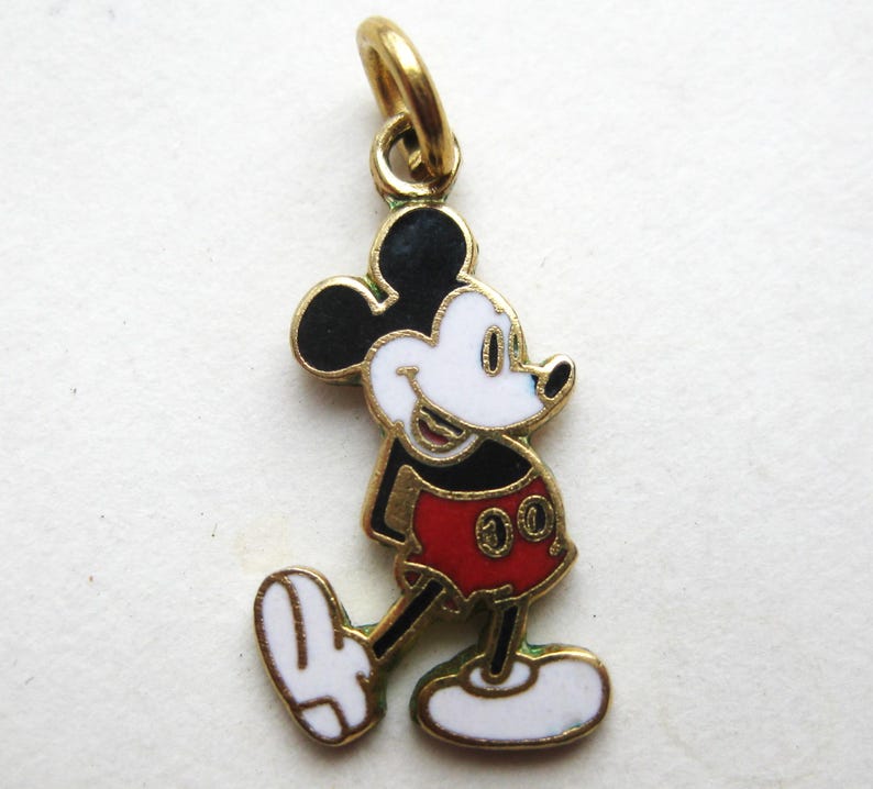 Puede incluir: Un charm de Mickey Mouse dorado con orejas negras, cara blanca y pantalones cortos rojos. El charm tiene un lazo dorado para sujetarlo a una pulsera o collar. El ic&oacute;nico personaje de dibujos animados se representa caminando.
