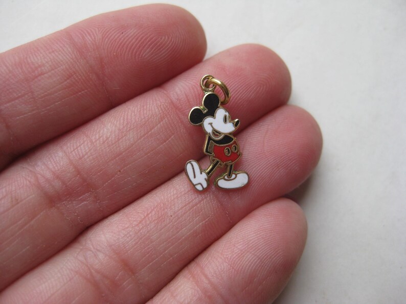 Puede incluir: Un peque&ntilde;o dije de Mickey Mouse de color dorado. El dije presenta una cabeza y orejas negras, una cara blanca, pantalones cortos rojos y guantes y zapatos blancos. El dije est&aacute; unido a un peque&ntilde;o lazo dorado.