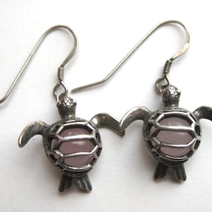 May include: A pair of silver-toned metal earrings featuring a turtle-shaped design. Each turtle has a cage-like shell structure that holds a smooth, round, pale pink stone in the center. The earrings are attached to simple silver-toned metal hooks.