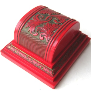 Op de afbeelding: Een felrode doos in antieke stijl met ingewikkelde bloemsnijwerken. De doos heeft een rechthoekige basis en een gebogen bovenkant, met goudkleurige accenten die het gedetailleerde ontwerp benadrukken. De algehele esthetiek is sierlijk en decoratief.